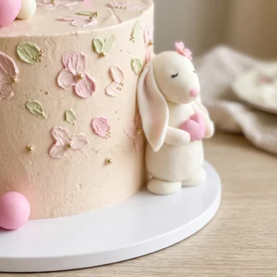 Boční detail dortu s bílým zajíčkem držícím růžové srdíčko, jemnými květinami a pastelovým narozeninovým zdobením.