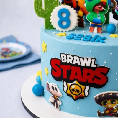Detail boku dortu Brawl Stars se světle modrým krémem, logem, hvězdičkami a plochými obrázky postaviček.