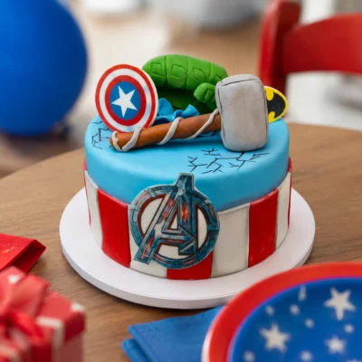 Modrý dort Avengers s červenobílým zdobením, plochým logem na boku a modelovanými superhrdinskými prvky na horní části.