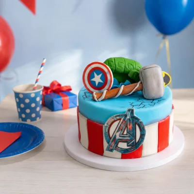 Narozeninový dort Avengers na slavnostním stole s modrými a červenými doplňky, balónky, dárkem a superhrdinskými dekoracemi.