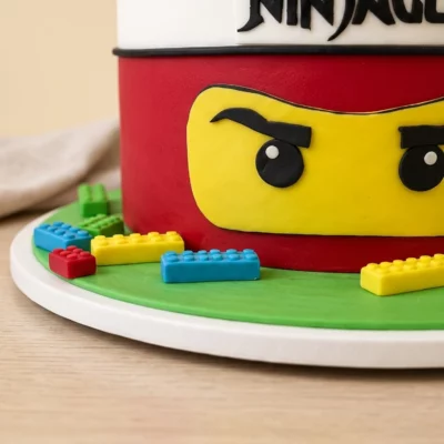 Detail boku dortu Ninjago se žlutou maskou, červeným potahem a barevnými LEGO kostičkami na zelené podložce.