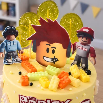 Horní detail dortu Roblox s hlavou se sluchátky, postavou v červené čepici a barevnými kostičkami.
